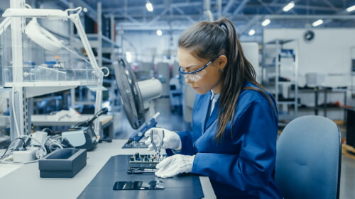 Young female with safety coat, safety glasses using plier to assemble printed circuit board for smartphone